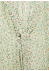 Blouse fleurie en tissu vert clair avec des fleurs blanches et beiges, un col en V et un détail noué à la taille.