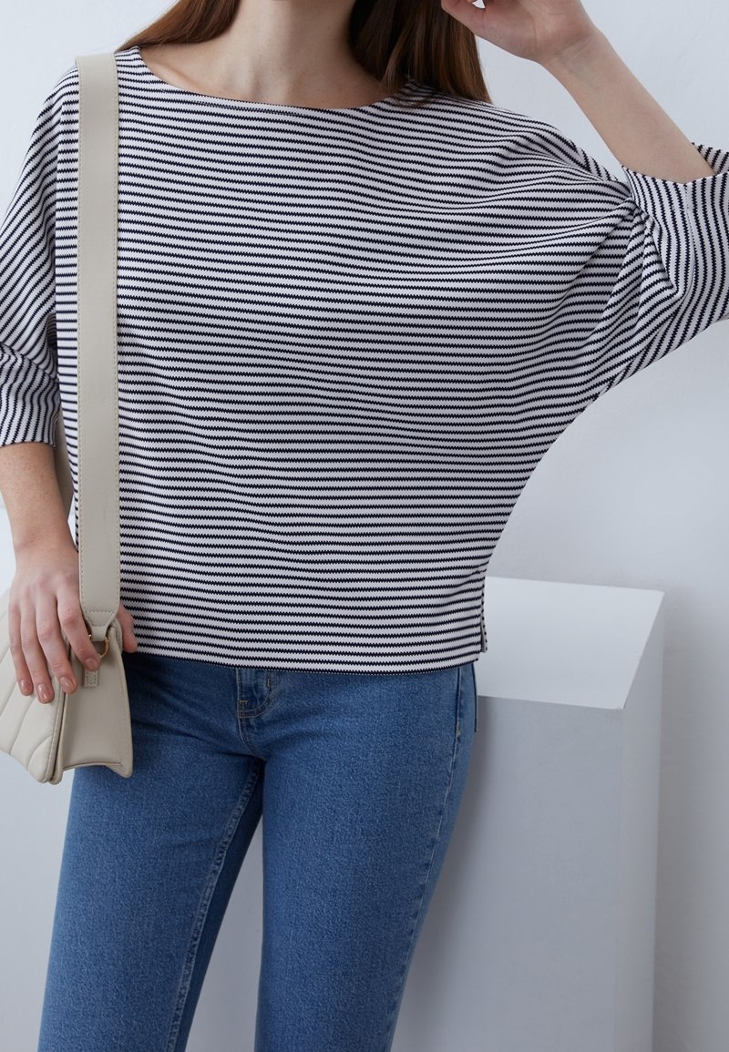 Femme portant un haut à manches chauve-souris rayé noir et blanc horizontalement, un jean bleu et un sac à bandoulière beige sur un fond clair uni.