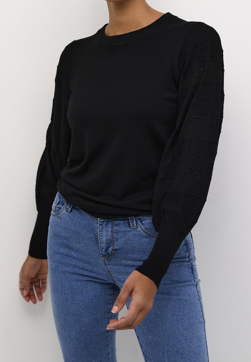 Svart tröja med texturerade ärmar, rund halsringning och ribbade manschetter, i kombination med blå skinny jeans. Tröjan har en mjuk, slät yta.