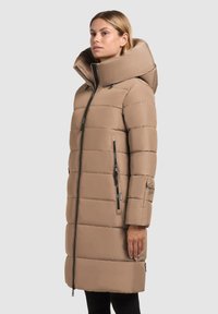 Lang beige quilted jakke med høj krave, vandret quiltet mønster, to sidelukninger og et logo-mærke på ærmet. Glat stoftekstur.