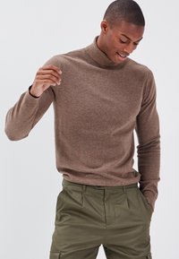 Brauner gestrickter Rollkragenpullover mit figurbetonter Passform und gerippten Bündchen, kombiniert mit olivgrünen Hosen. Weiche Textur, glatte Verarbeitung.