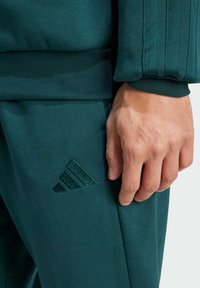 Sweatshirt et pantalon Adidas verts en tissu doux ; présente un logo texturé sur le pantalon et un détail de poignet côtelé sur le sweatshirt.