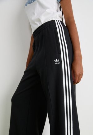 Zbliżenie osoby noszącej czarne spodnie dresowe Adidas z białymi paskami i logo, zestawione z białą koszulką.