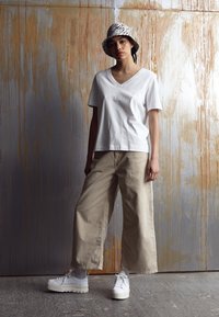 T-shirt bianco a V, pantaloni beige a gamba larga e sneakers bianche con plateau. Un cappello a secchiello a righe bianche e nere completa il look su uno sfondo testurizzato.