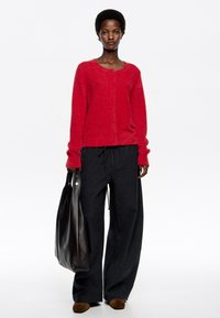 Cárdigan rojo de felpa con cuello redondo y cierre de botones, combinado con pantalones sueltos de color gris oscuro y un gran bolso de piel negro.