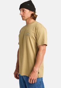 Man in een zwarte gebreide beanie, beige Billabong t-shirt en blauwe jeans, staande in profiel tegen een effen witte achtergrond.
