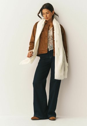 Hnědá semišová bunda přes bílý krajkový top, tmavé zvonové džíny a bílý šátek se zlatou výšivkou. Oblečení doplňují hnědé boty.