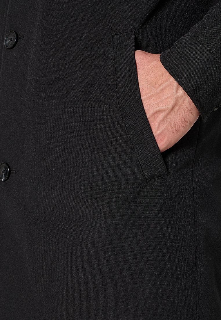 Zwarte jas met een close-up van een hand in een zijzak. De stof lijkt glad met een gestructureerde afwerking en heeft zwarte knopen.