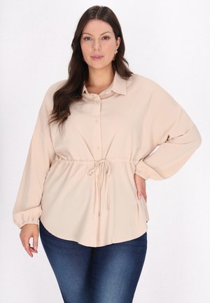 Beige knap-ned skjorte med lange ærmer, snor i taljen og elastiske manchetter; glat tekstur med en afrundet kant. Båret med blå jeans.