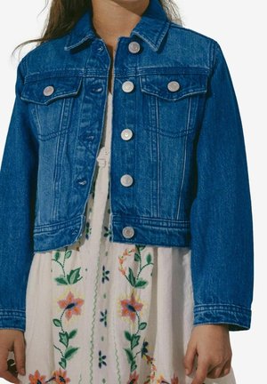 Bambino che indossa una giacca di jeans blu sopra un vestito bianco con ricamo floreale colorato, in piedi con le braccia rilassate lungo i fianchi.