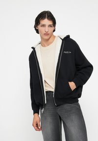 Hoodie noir zippé avec doublure en shearling blanche et logo brodé. Associé à un t-shirt beige et un jean gris. Coupe décontractée.