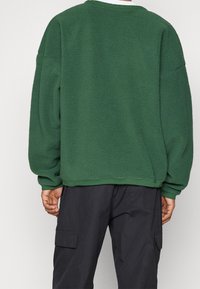 Zelená fleecová mikina s texturovaným povrchem, spuštěnými ramena a žebrovaným výstřihem, v kombinaci s tmavými cargo kalhotami.