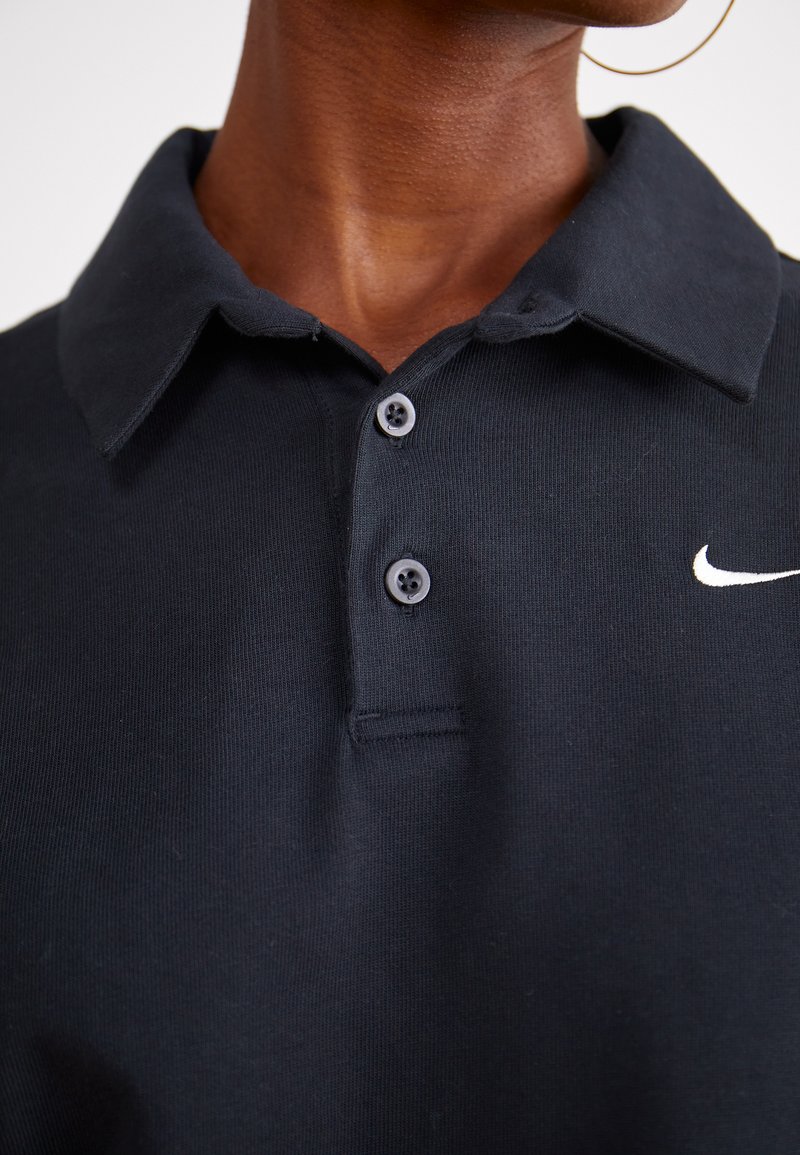 Polo negro de algodón con cuello abotonado, presenta dos botones grises y un logo blanco de Nike en la parte izquierda del pecho.