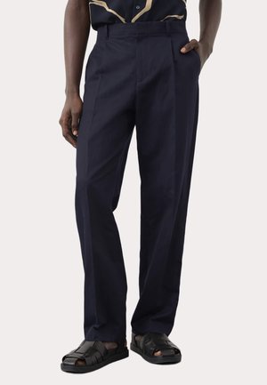 Homme portant un pantalon tailleur bleu marine foncé, des sandales en cuir tressé noir et une vue partielle d'une chemise noire et beige.