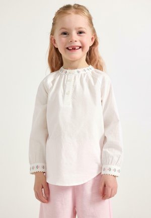 Młoda dziewczyna uśmiechająca się, ubrana w białą bluzkę z długim rękawem z kwiatowym haftem na kołnierzyku i mankietach oraz jasnoróżowe spodnie.