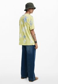Camisa de manga corta amarillo claro con patrón floral azul, combinada con jeans de mezclilla azules de corte holgado y zapatos slip-on color crema. Sombrero de cubo verde oscuro.