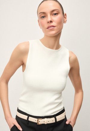 Mujer con el cabello hacia atrás, vestida con una blusa blanca sin mangas y pantalones negros con un cinturón beige claro, de pie con las manos en los bolsillos.