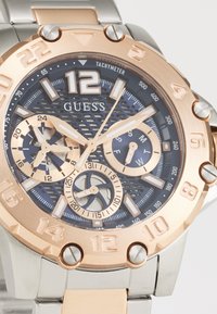 Montre analogue en argent et or rose avec un cadran bleu texturé, grand chiffre 12, sous-cadrans chronographes et bracelet en acier inoxydable.