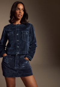 Niebieska granatowa zamszowa kurtka z zapięciem na guziki z przodu i dwoma kieszeniami na piersi, w zestawie z pasującą spódnicą o gładkiej fakturze i bocznych kieszeniach.