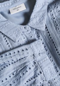 Světle modrá blůza s bohatou výšivkou, která má výřezové vzory, zapínání na knoflíky a špičatý límec. Vyrobena z texturované tkaniny.