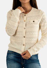 Donna che indossa un cardigan a maglia color crema con un motivo a diamante e cuore, abbottonato sul davanti, e jeans blu.