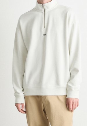 Weißer Pullover mit halbem Reißverschluss und schwarzem "HUGO"-Logo auf der Brust, getragen von einer Person in hellbrauner Hose, vor einem schlichten weißen Hintergrund stehend.