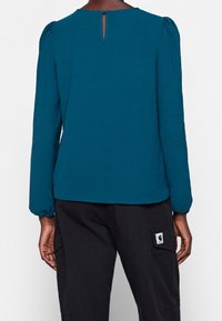 Maglia lunga a maniche con spalle a sbuffo in teal, apertura a chiave sul colletto e tessuto strutturato. Abbinata a pantaloni cargo neri con tasca laterale.