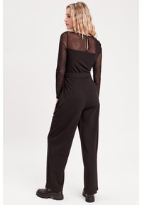 Haut à manches longues noir en tissu transparent à pois, associé à un pantalon noir taille haute. Le mannequin fait face à droite, mettant en valeur les détails du dos et les chaussures.