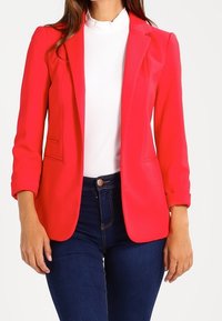 Blazer rouge en tissu texturé, avec des revers à cran et deux poches avant, associé à un haut blanc et un jean bleu foncé.