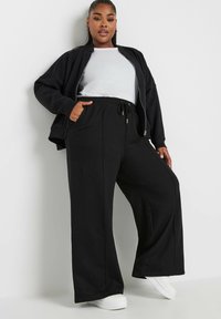 Svart zip-up bomberjacka över en vit t-shirt, i kombination med löst sittande svarta sweatpants. Byxorna har elastisk midja och sidofickor.