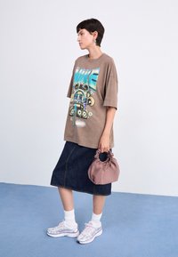 Person med kort, mörkt hår som bär en överdimensionerad brun t-shirt med tryck, mörk denimkjol, vita strumpor, silverfärgade sneakers och håller en liten mauve handväska.