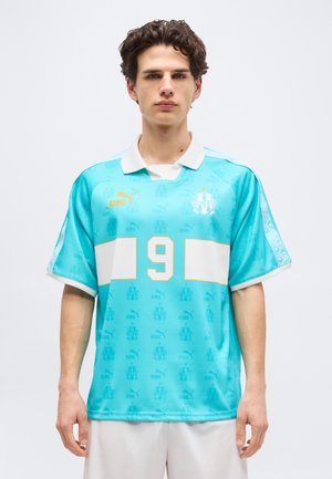 Νέος άνδρας που φοράει αθλητική φανέλα σε τιρκουάζ και λευκό με τον αριθμό 9 και το λογότυπο Puma, στέκεται μπροστά σε απλό φόντο.