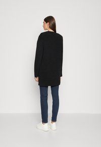 Lang sort sweater lavet af et struktureret stof, med en afslappet pasform og sænkede skuldre, parret med mørkeblå jeans og hvide sneakers.