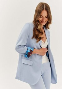 Svetle modrý blejzr s květinovým vzorem na manžetách, pohodlný střih, dvě přední kapsy a hladká textura, nošený přes krémový vršek.