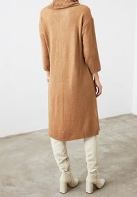 Robe col roulé oversize côtelée en tissu tricot camel ; longueur mi-mollet avec une silhouette droite et des bottes crème ceinturées.