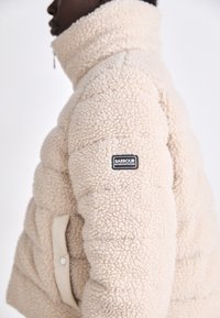 Beez värvusega pehme puffer jakk, mis on valmistatud pehmest ja tekstuuriga kangast, kõrge krae, nööbiga tasku ja must logopärastik käisel.