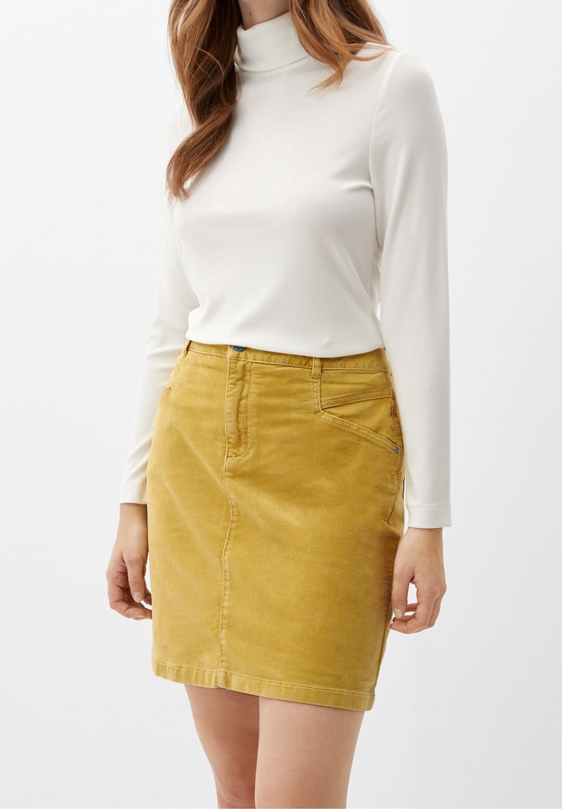 Vit långärmad turtleneck-top i kombination med en gul kordflodds-kjol, som har en framficka och en subtil struktur.