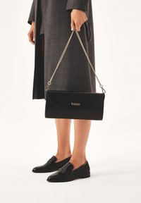 Zwarte leren clutch met een kettinghengsel, voorzien van een gebogen flap, een merklogo en een soepele textuur, vastgehouden door een persoon in een grijze jas.