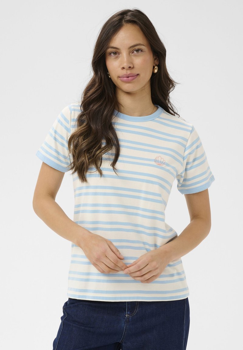 Jeune femme aux longs cheveux bruns portant un t-shirt à rayures bleues clair et blanches avec un petit motif de coquillage sur la poitrine, et un jean foncé.
