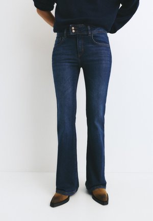 Oseba, ki nosi temno modre široke kavbojke, rjave koničaste čevlje in temno dolg rokav na enobarvnem belem ozadju.