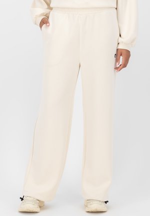 Cremefarbene, weit geschnittene Sweatpants aus weichem Stoff mit elastischem Bund, seitlichen Taschen und einem kleinen Logo-Patch auf der linken Seite.