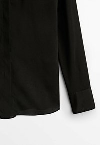Chemise noire à manches longues avec poignets pointus et ourlet incurvé, présentée sur fond blanc.