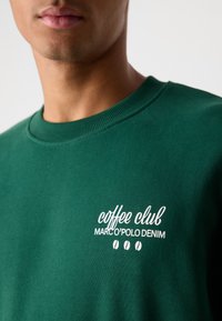 Groene sweatshirt met een ronde hals, voorzien van witte tekst: "coffee club MARCO'POLO DENIM" en drie koffieboon grafieken. Zachte textuur.