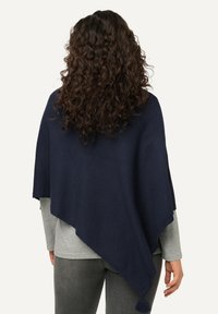 Navy sininen neulottu poncho, jossa on kolmiomainen muotoilu, viimeistelty tupsuilla. Vaate on yhdistetty harmaan pitkähihaisen paidan kanssa, luoden rentoa ja kerroksellista ilmettä.
