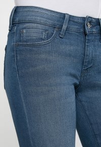 Jeans in denim lavaggio blu medio, con un design aderente, stile a cinque tasche e dettagli in metallo nella cintura e nelle tasche.