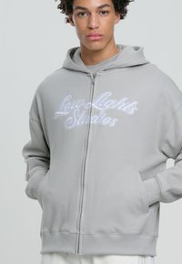 Felpa grigia con zip realizzata in morbido tessuto, con un logo ricamato "Low Lights Studios" in bianco, tasche a marsupio e polsini a costine.
