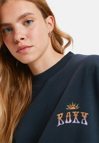 Granatowy sweter z ribbowanym dekoltem, z multikolorowym haftem "ROXY" oraz grafiką słońca na klatce piersiowej. Miękki, bawełniany materiał.