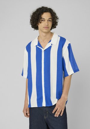 Jeune homme aux cheveux bouclés portant une chemise boutonnée à manches courtes à rayures verticales bleues et blanches et un pantalon foncé.