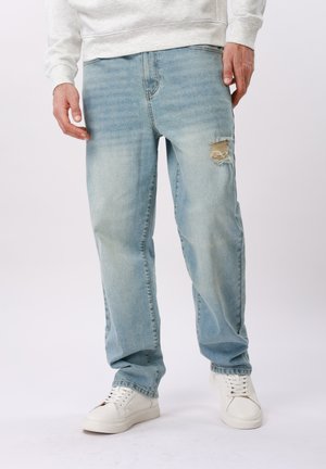 Homme portant un jean bleu clair déchiré et des baskets blanches, debout les mains détendues le long du corps sur un fond blanc.