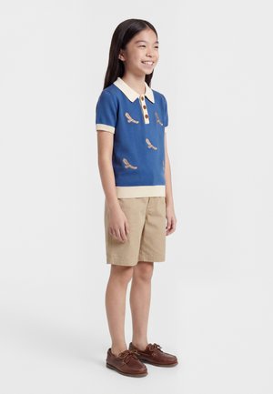 Niña sonriente, vistiendo un polo azul de manga corta con estampados de pájaros, pantalones cortos beige y zapatos náuticos marrones, de pie contra un fondo liso.